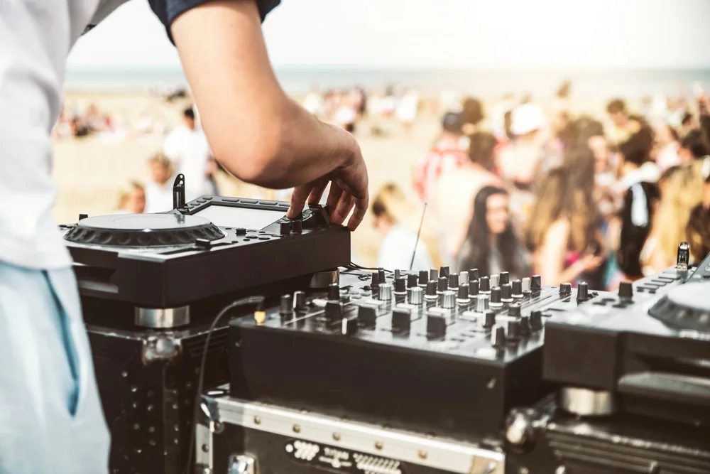 A DJ’s hands adjusting a mixer as a crowd dances in the background